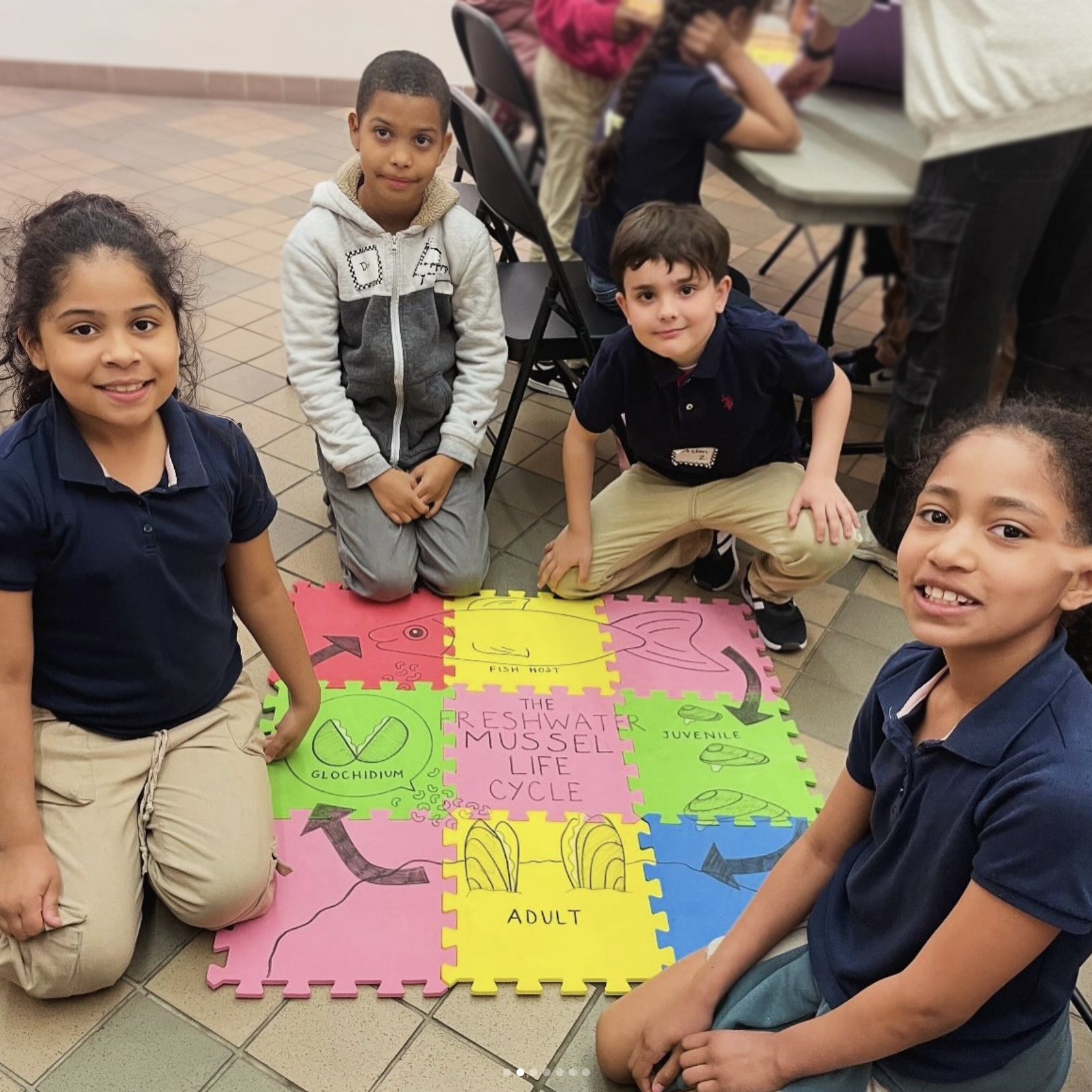 Ethan Allen School Students Explore the Freshwater Mussel Life Cycle Four Ethan Allen School students sit on the floor around a colorful puzzle mat showing the freshwater mussel life cycle, smiling as they engage in the learning activity.